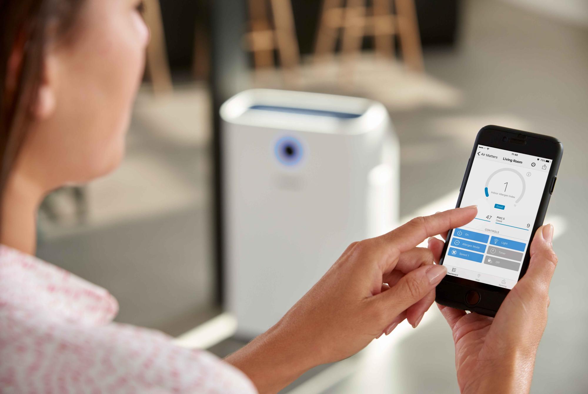 Persona izmanto viedtālruņa lietotni, lai kontrolētu baltu Philips Climate Care 2in1 gaisa attīrītāju, demonstrējot viedās vadības, gaisa attīrīšanas un eleganta dizaina funkcijas.