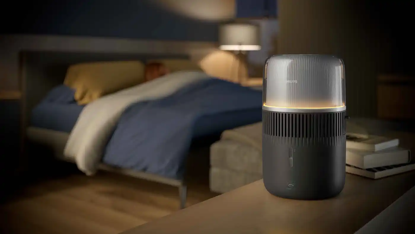 Air-Humidifier-3-fi Philips gaisa mitrinātājs uz naktsgaldiņa maigi apgaismotā guļamistabā, izceļot Philips gaisa mitrinātāju līnijas miegu uzlabojošās priekšrocības ar tā eleganto dizainu un izcilo mitrināšanas tehnoloģiju.