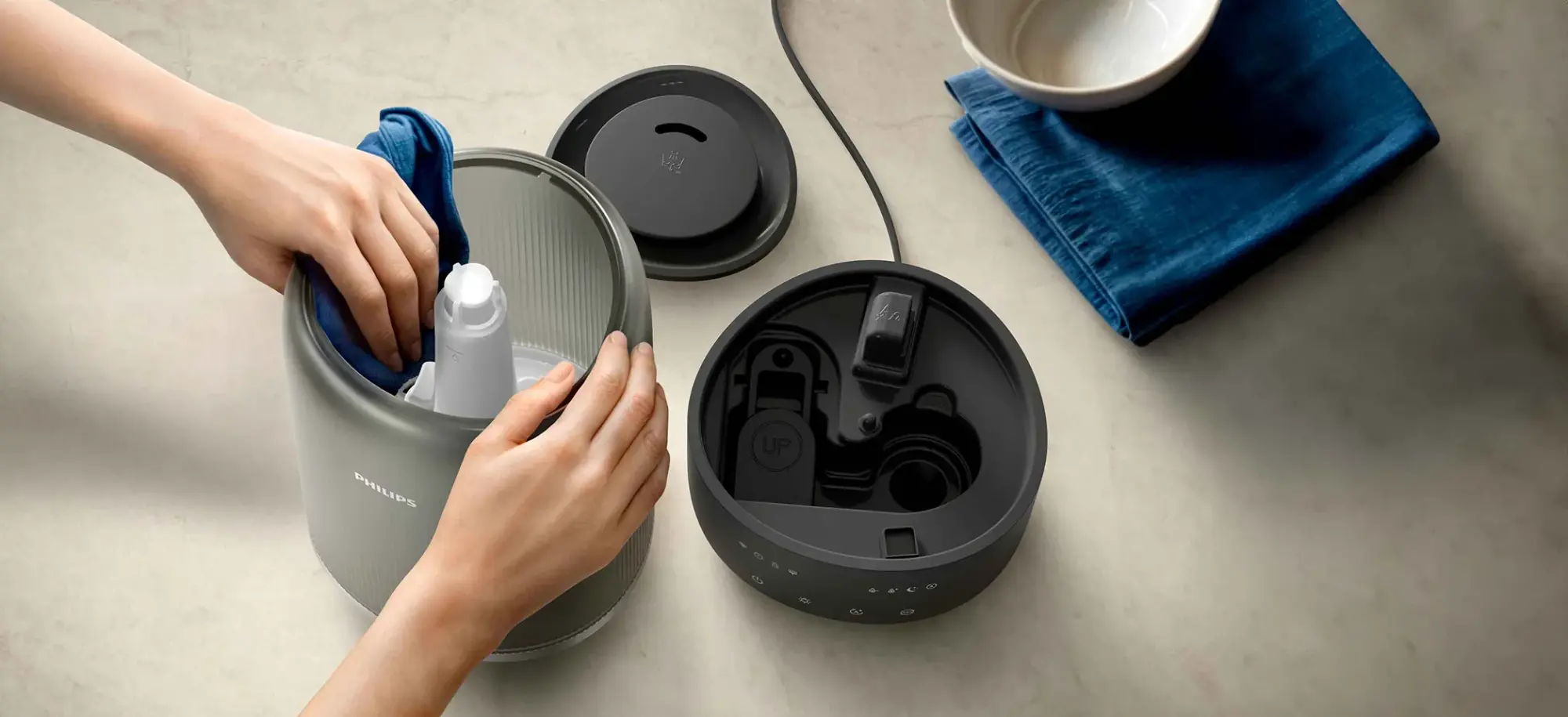 Air-Humidifier-6-fi Rokas tīra atvērtu Philips gaisa mitrinātāju, izceļot tā vienkāršo apkopi. Redzama gaisa mitrinātāja tvertne, pamatne, vāks un iekšējās daļas.