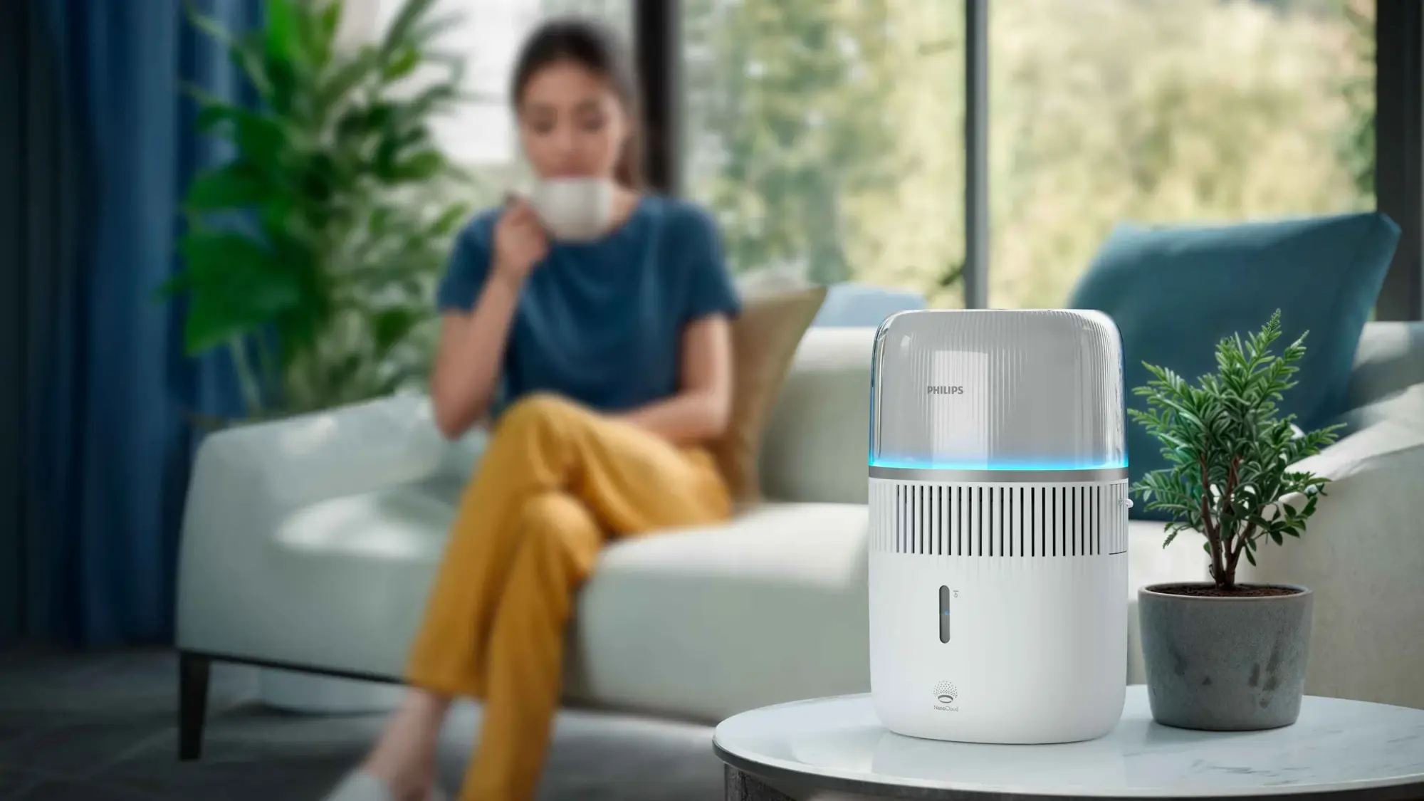 Air-Humidifier-8-fi Philips gaisa mitrinātājs uz galda, izstaro maigu zilu mirdzumu, blakus podā audzētam augam, sieviete malko tēju. Philips gaisa mitrinātājs: izcils dizains, nepārspējama gaisa kvalitāte.