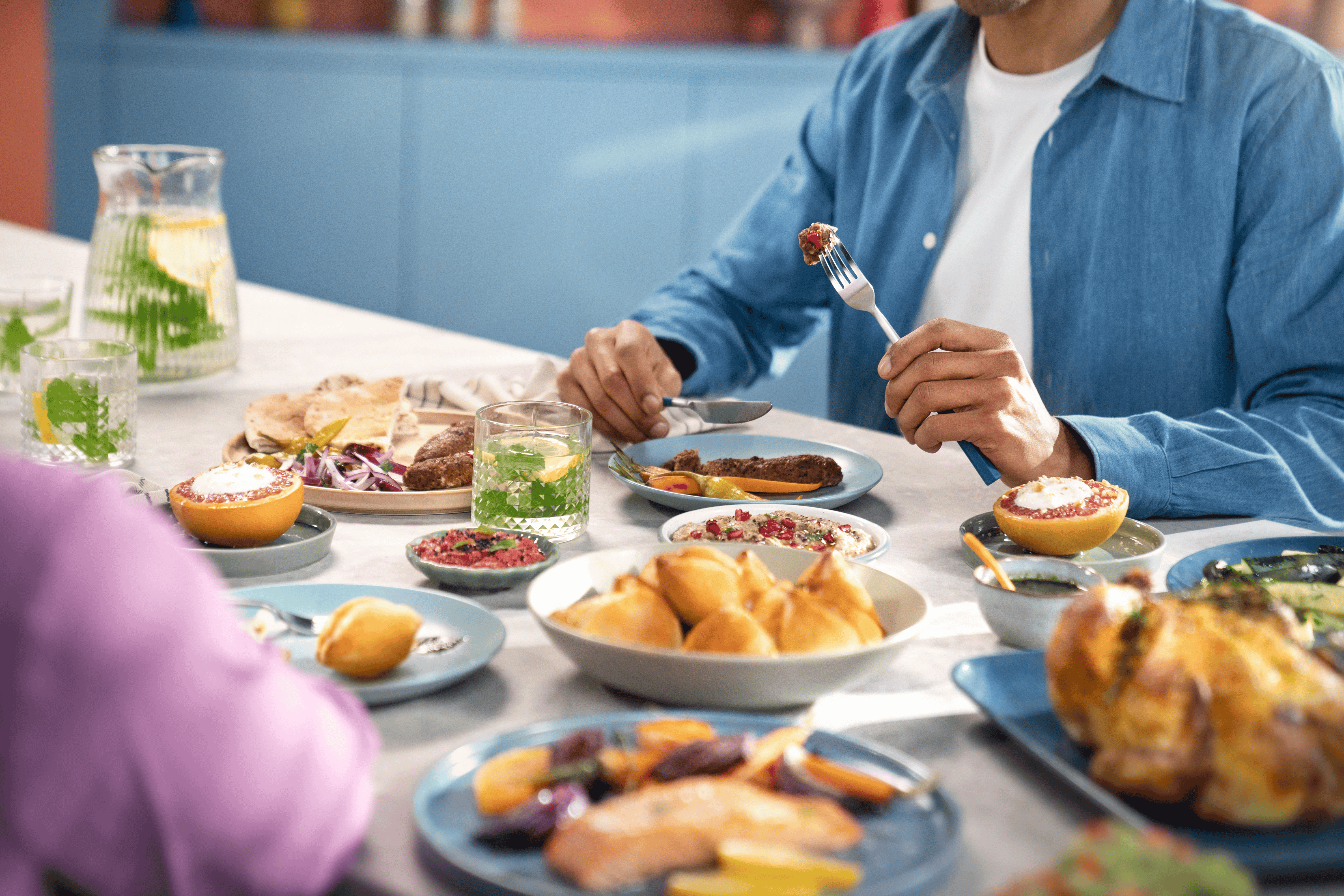 Cilvēki bauda maltīti pie galda, kas piepildīts ar dažādiem krāsainiem ēdieniem un atspirdzinošiem dzērieniem.