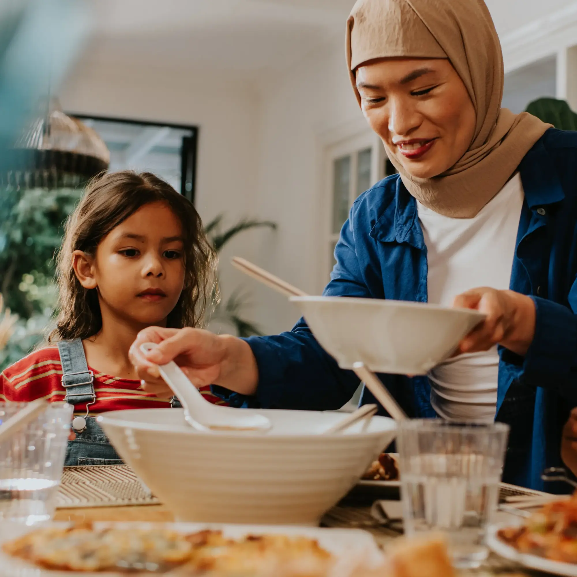 Sieviete pasniedz ēdienu bērnam pie pusdienu galda, radot siltu un ģimenisku atmosfēru.