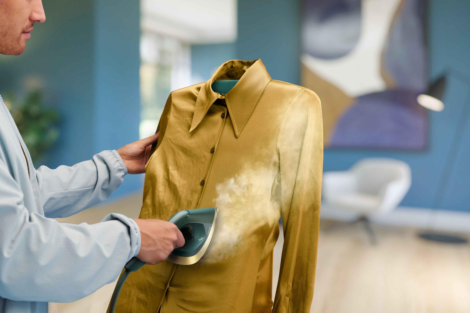 Philips universālā All-in-One apģērbu kopšanas ierīce: persona, kas izmanto tvaicētāju zelta krāsas apģērbam, demonstrē tūlītēju grumbu likvidēšanu ar tvaiku. Moderna istaba.