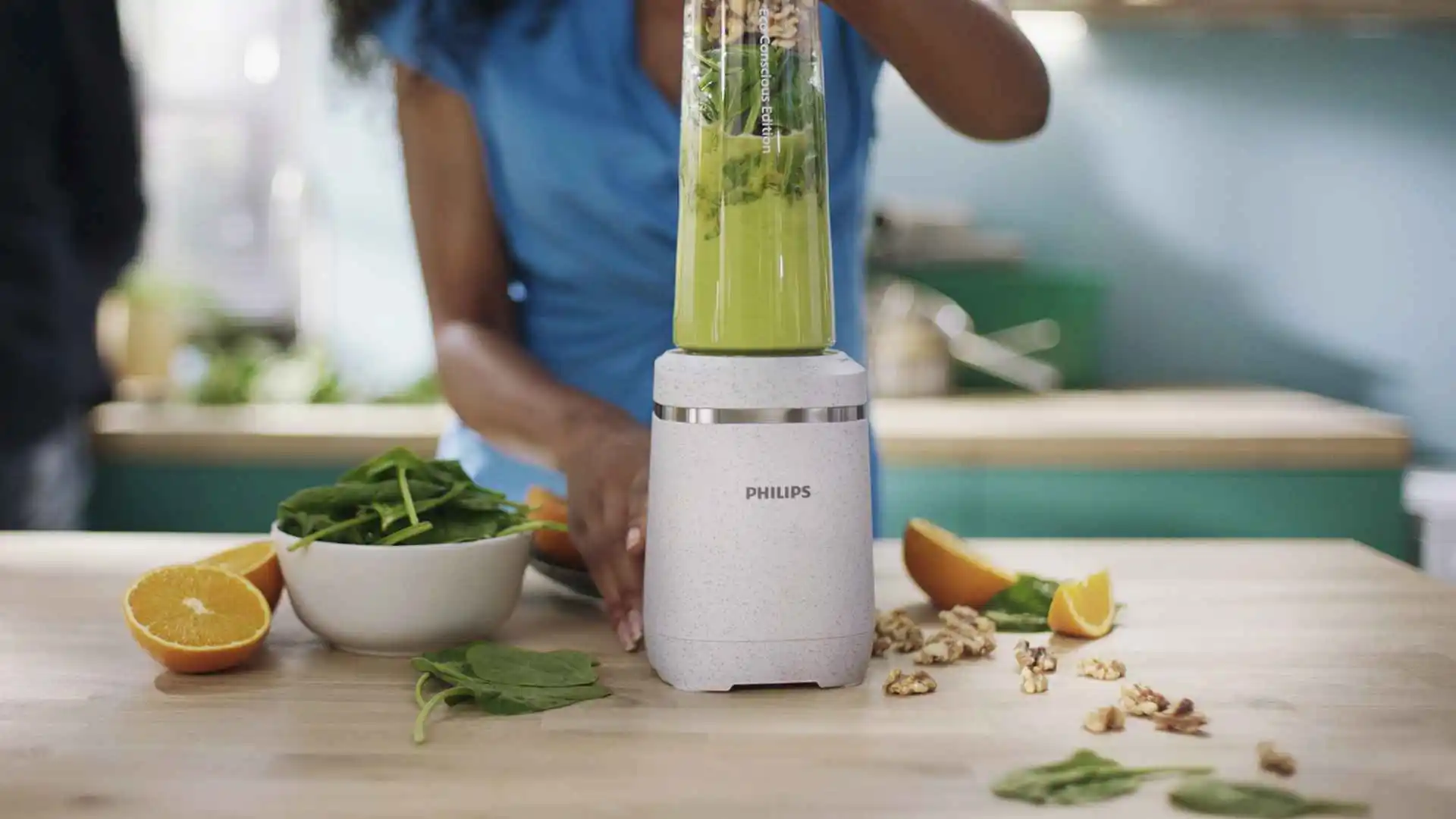 Sieviete gatavo zaļo kokteili ar Philips Blend & Juice virtuves portatīvo blenderi; spināti, apelsīni un valrieksti uz koka letes demonstrē lietotāja pieredzi.