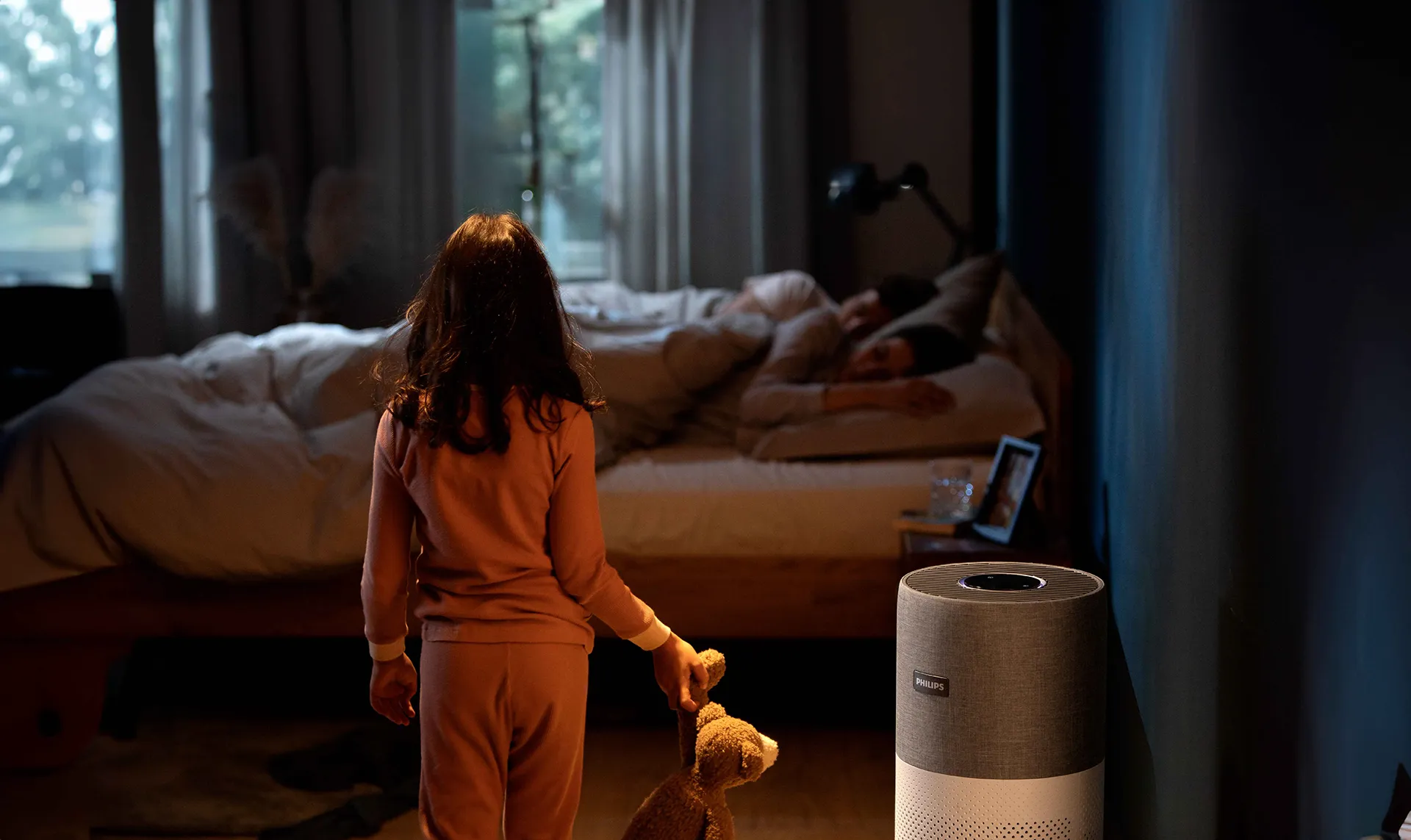 Philips Climate Care gaisa attīrītājs nodrošina tīru gaisu guļošai ģimenei. Bērns ar lācīti stāv pie attīrītāja elegantā, cilindriskā dizaina vāji apgaismotā guļamistabā.