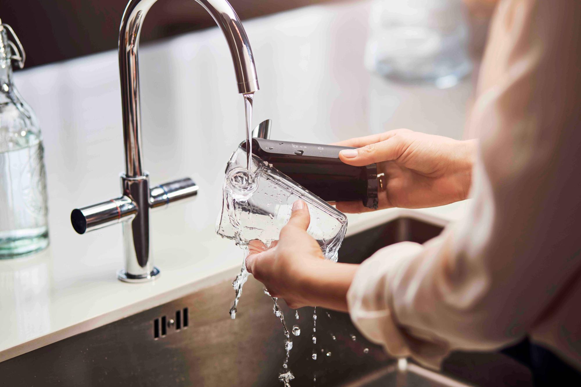 Coffee-19-fi Cilvēks pēc lietošanas skalo Philips kafijas automāta tīrā ūdens tvertni pie izlietnes, uzsverot vienkāršo tīrīšanas funkciju. Tas nodrošina svaigu kafijas garšu katru reizi. Redzams spīdīgs krāns.