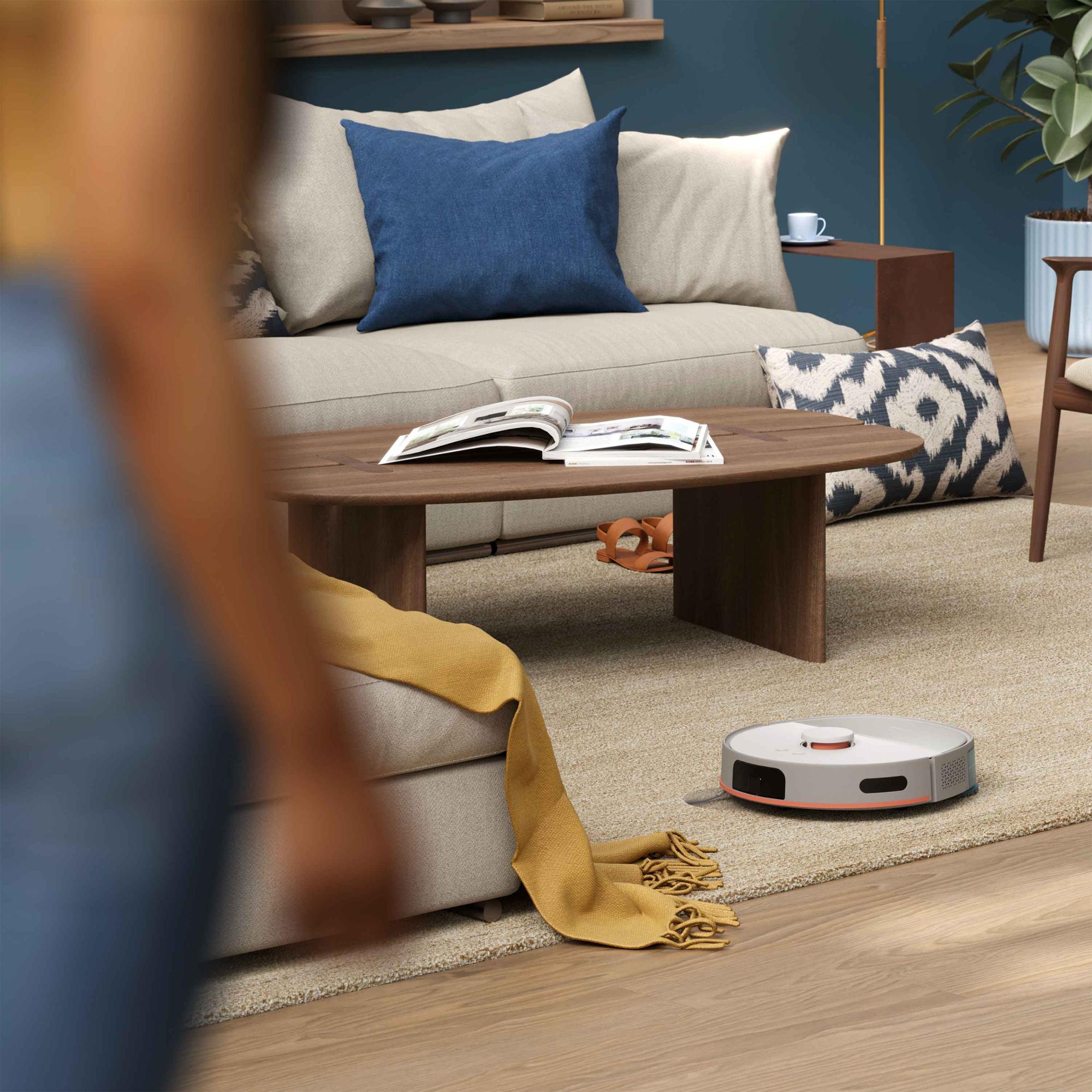 Philips Floor Care robotizētais putekļsūcējs pārvietojas ap mēbelēm uz smilškrāsas paklāja, demonstrējot savus viedos sensorus un efektīvu tīrīšanu modernā mājā.