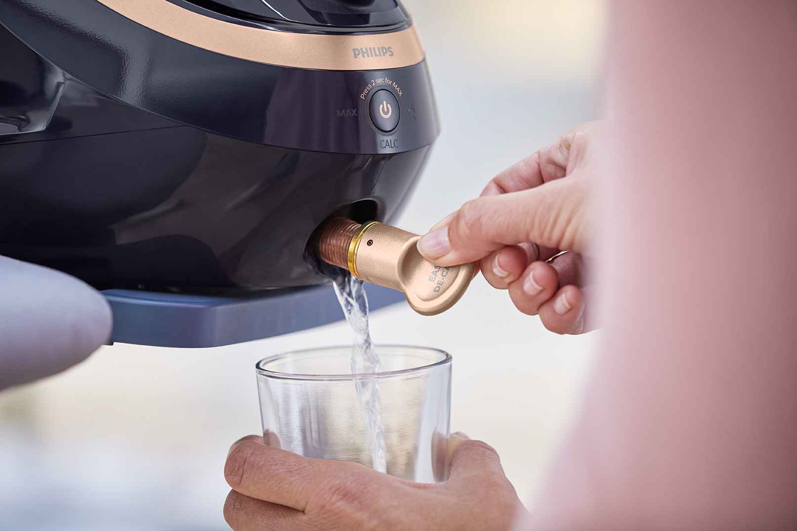 Tuvplānā redzama Philips Garment Care tvaika gludināšanas sistēmas Easy De-Calc funkcija. Ūdens notek glāzē, demonstrējot ierīces ērto un vienkāršo apkopes dizainu.