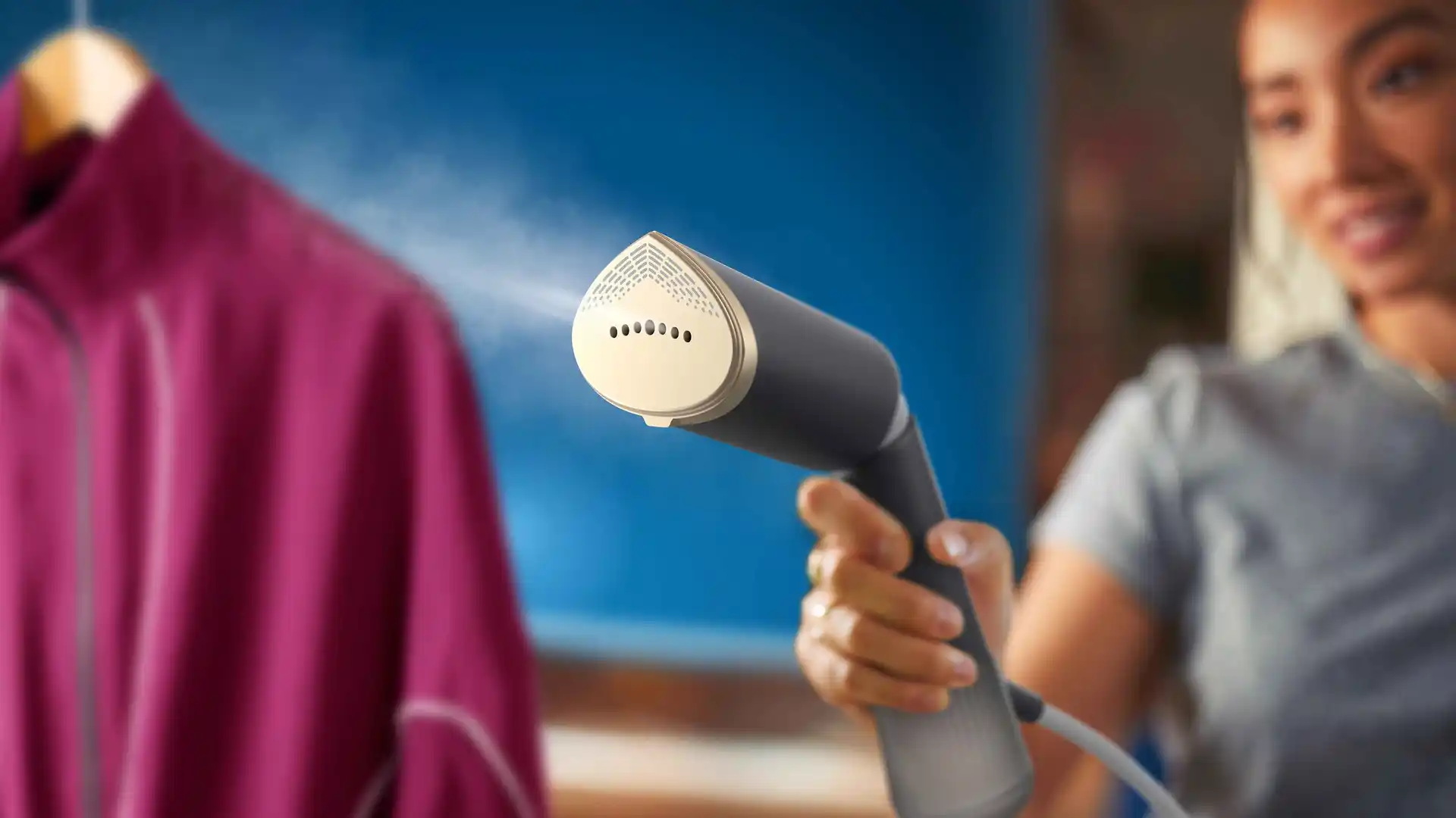 Persona lieto Philips rokas apģērba tvaicētāju uz fuksīna krāsas jakas. Rokas dizains ar redzamu tvaiku demonstrē tās efektīvu grumbu likvidēšanu.