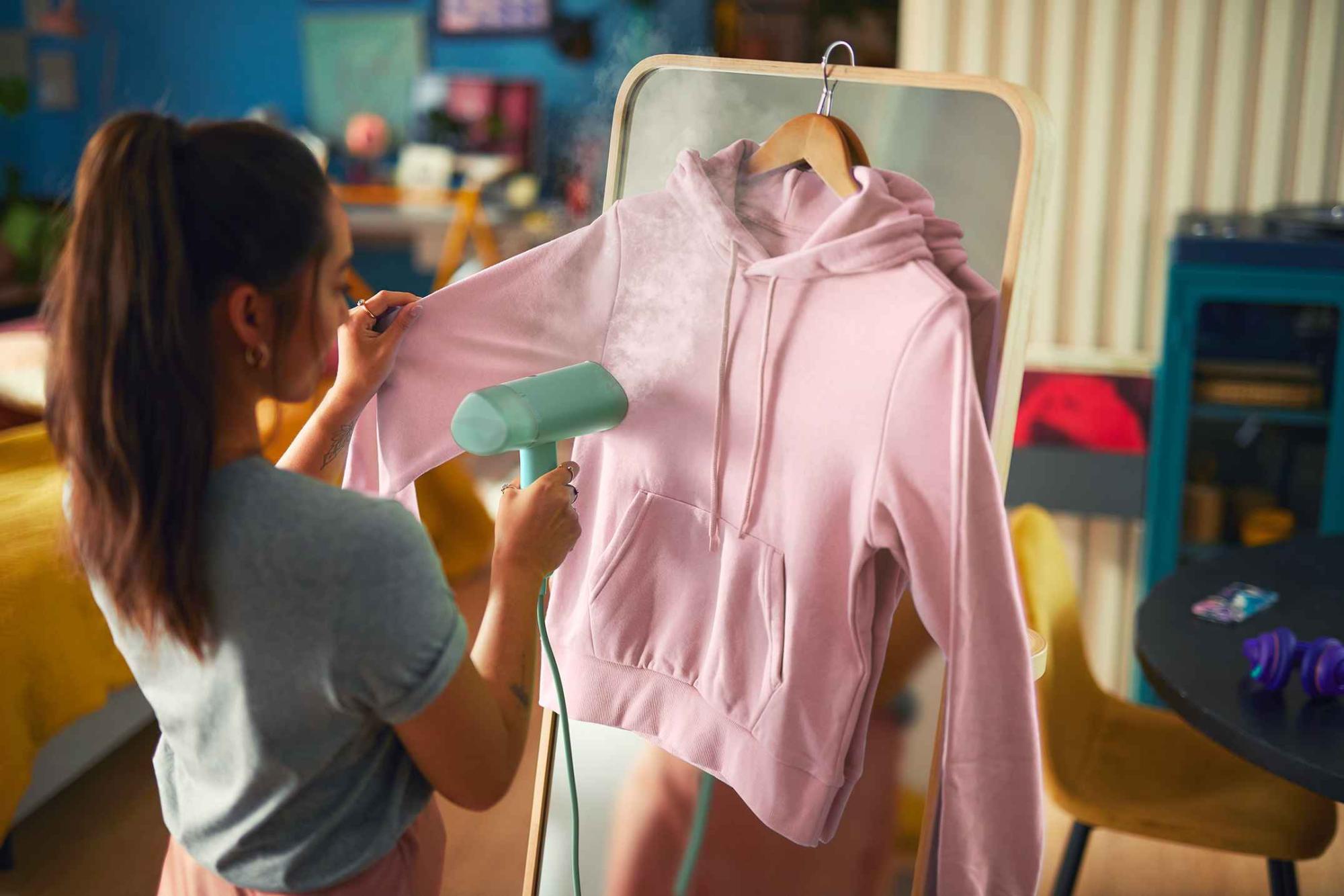 Philips apģērbu kopšana: Sieviete tvaicē rozā kapučjaku ar rokas apģērbu tvaicētāju, tvaiks ceļas augšup, grumbas izzūd, redzeslokā pakaramais un spogulis.