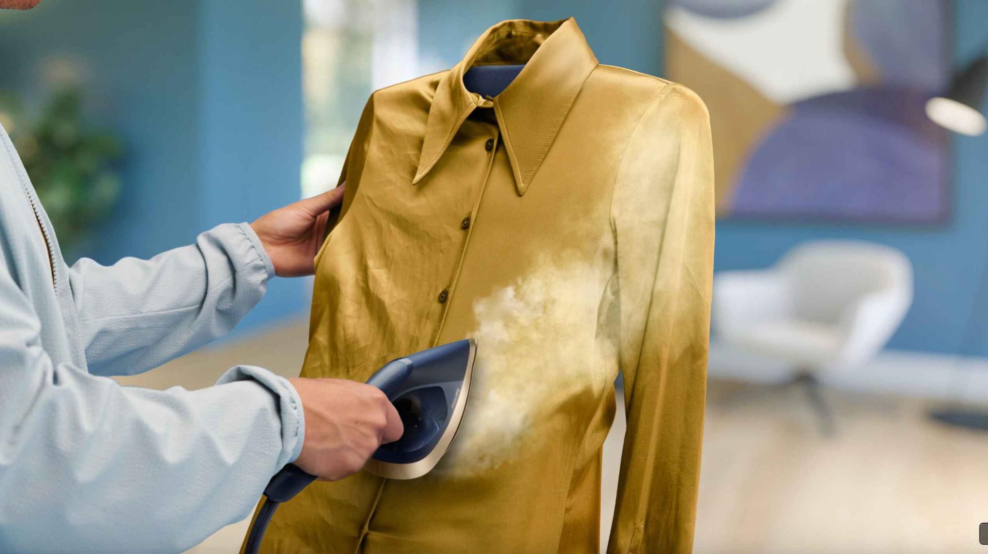 Philips apģērbu kopšanas tvaicētājs darbībā, ātri izlīdzinot zelta satīna kreklu; uzsver vieglu grumbu likvidēšanu un auduma kopšanu.