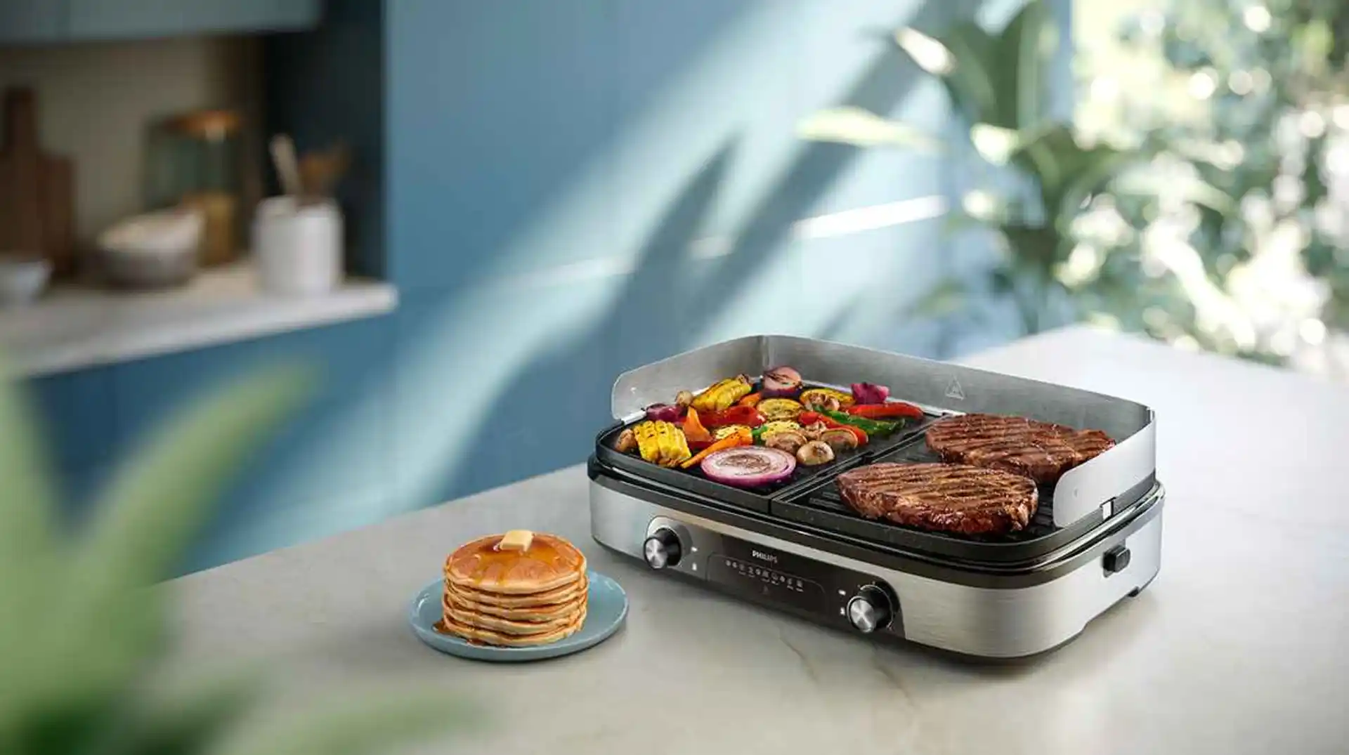 Philips grilu un tosteru līnija: augstas klases elektriskais grils, kas meistarīgi apcep steikus un grilē spilgtas krāsas dārzeņus, ko papildina pankūku kaudze modernā virtuvē.