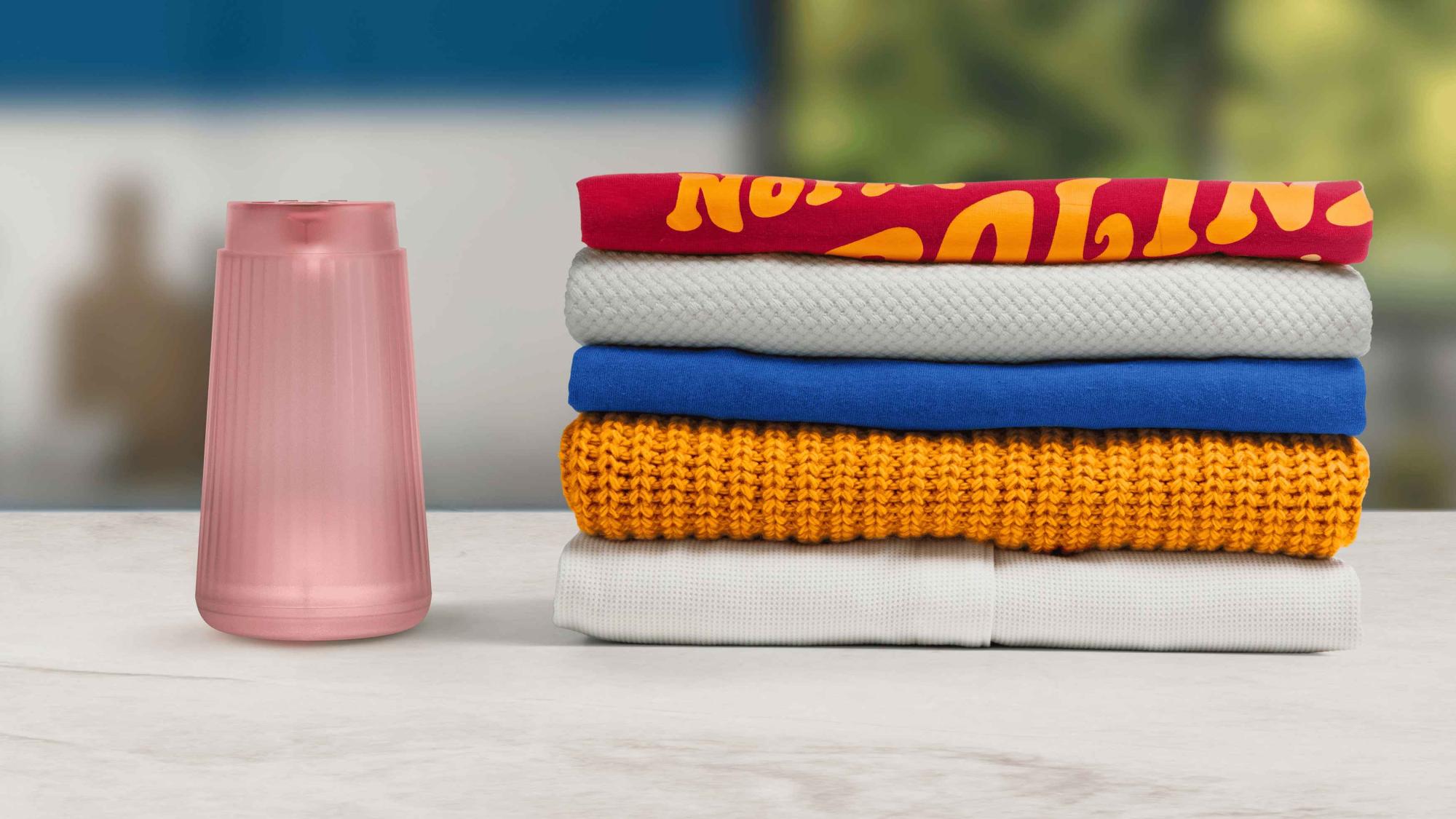 Philips rokas apģērba tvaicētāji: rozā ierīce, kas attēlota kopā ar drēbju kaudzi. Nodrošina ātru apģērba kopšanu un ērtu kopšanu mājās vai ceļojumā.