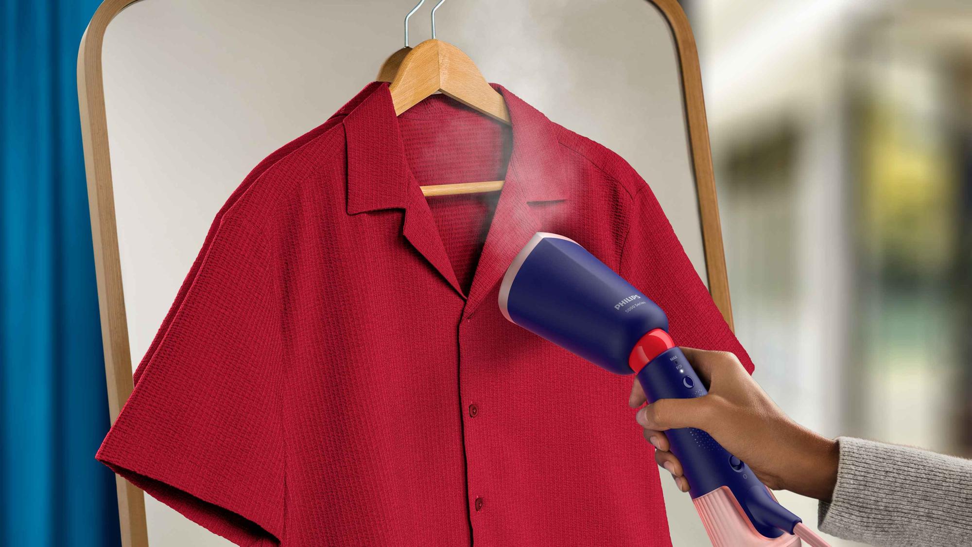 Philips rokas apģērba tvaicētāji bez piepūles iztīra grumbas no sarkana krekla. Ergonomiskais dizains ar ziliem un rozā akcentiem ērtai lietošanai.