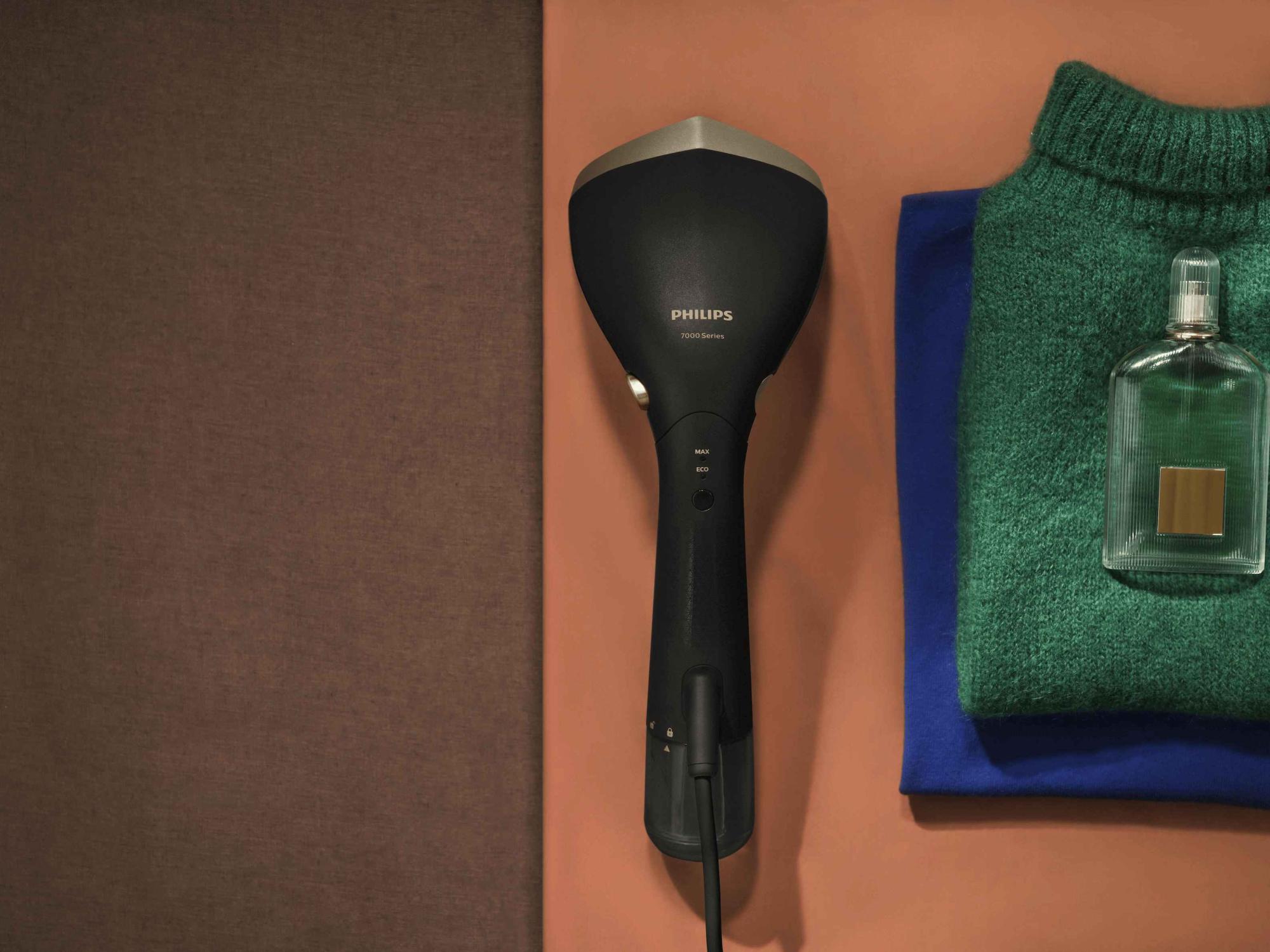 Studijas attēls ar elegantu, melnu Philips rokas apģērbu tvaicētājuno Philips Garment Care līnijas, kas novietots kopā ar salocītiem džemperiem un smaržu pudelīti uz silti brūnas virsmas.