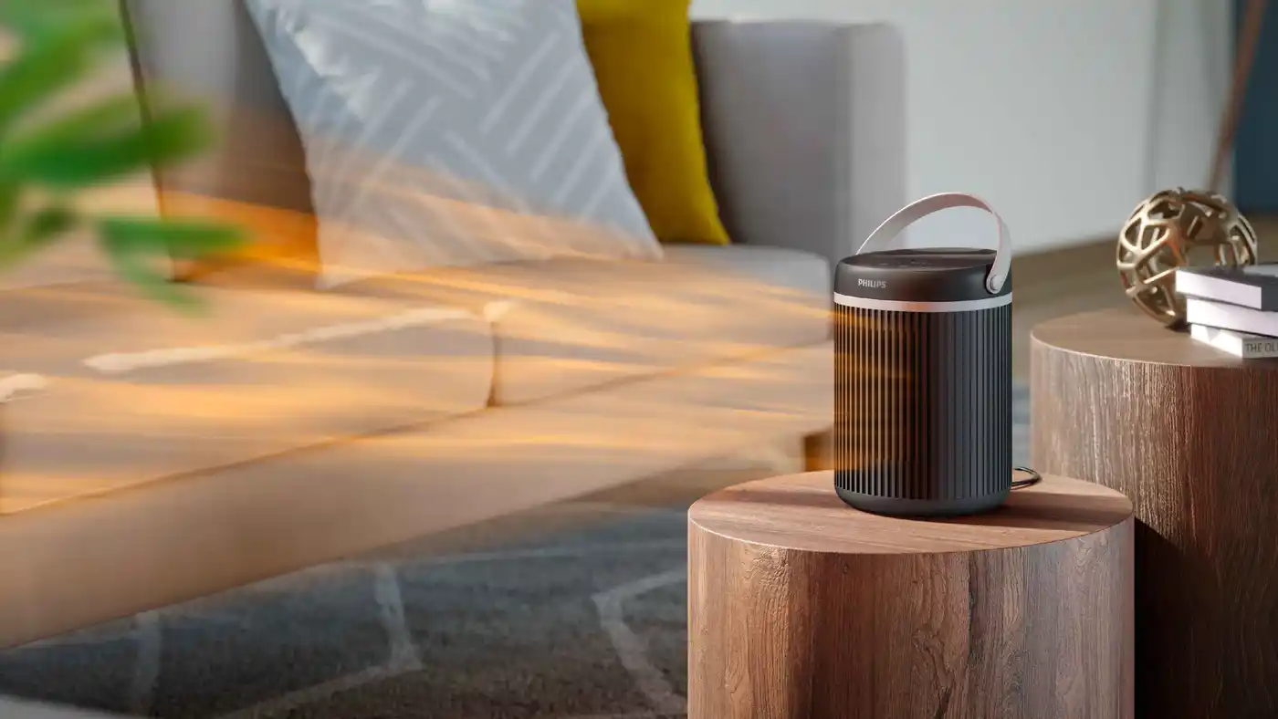 Philips sildītājs: augstākās klases Climate Care sildītājs ar rokturi un elegantu dizainu, kas novietots uz koka galda un nodrošina siltumu pie dīvāna labi apgaismotā dzīvojamā telpā.