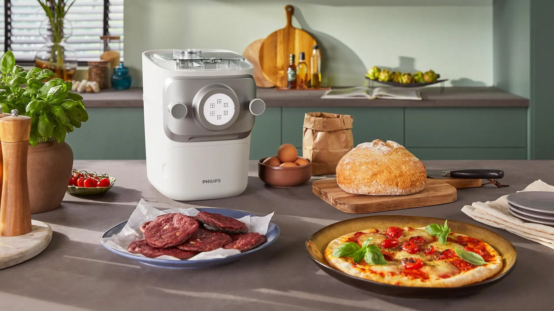 Attēlā redzama virtuves lete ar Philips virtuves ierīci - Philips pastas gatavotāju, maizes klaipu, olām, gaļas šķēlītēm uz šķīvja un picu. Fonā redzami virtuves piederumi, garšvielas un dārzeņi.