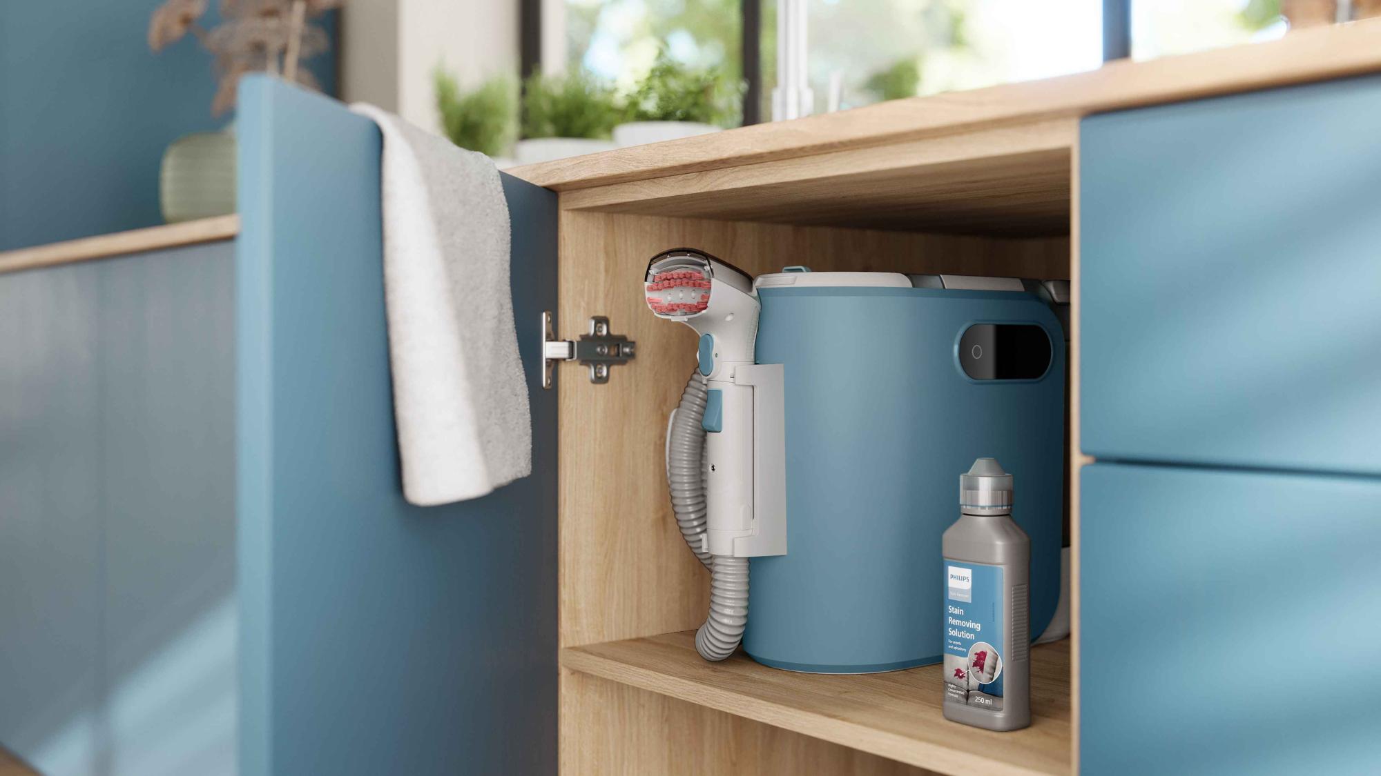Attēlā redzams Philips traipu tīrītājs ar birsti, šļūteni un traipu tīrīšanas līdzekli, kas glīti ievietots gaiša koka skapītī. Philips plankumu tīrītājs: nepārspējama tīrīšanas jauda kompaktā dizainā.