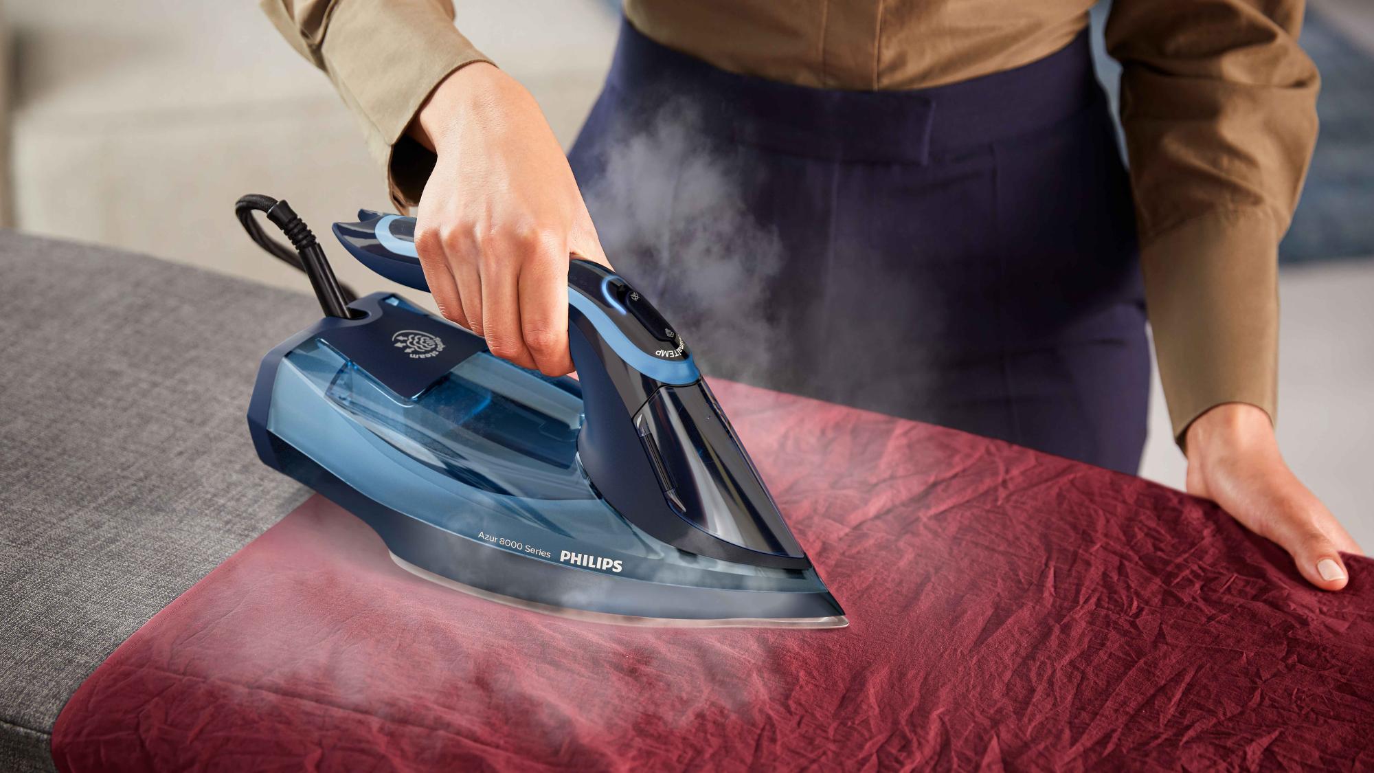 Philips tvaika gludekļi: Cilvēks gludina sarkanu audumu ar zili melno Azur 8000 sērijas tvaika gludekli, izdalot tvaiku, demonstrējot gludināšanas precizitāti.