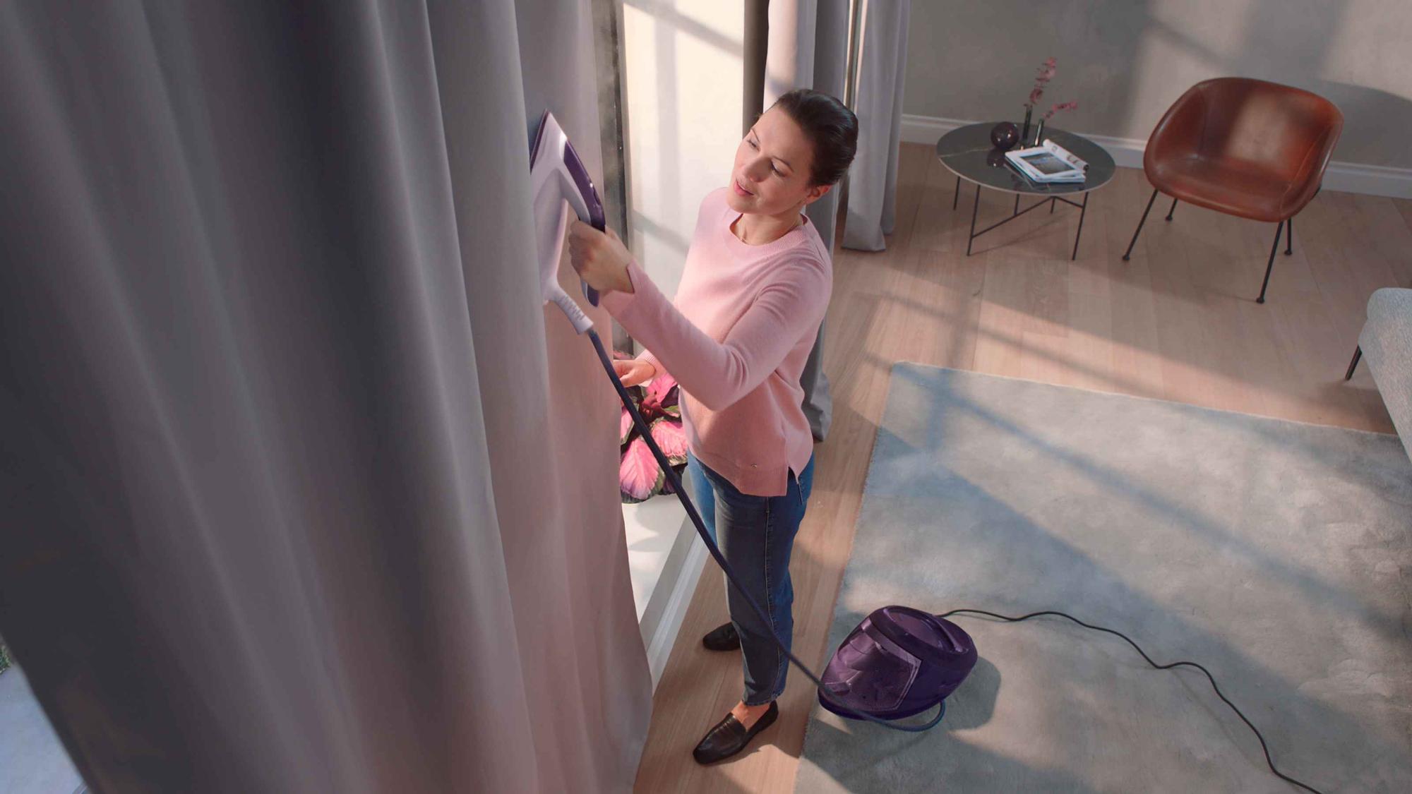 Sieviete tīra aizkarus ar Philips gludināšanas sistēmu, demonstrējot tā lietošanas ērtumu. Tiek izcelta jaudīgā grumbu izlīdzināšana.