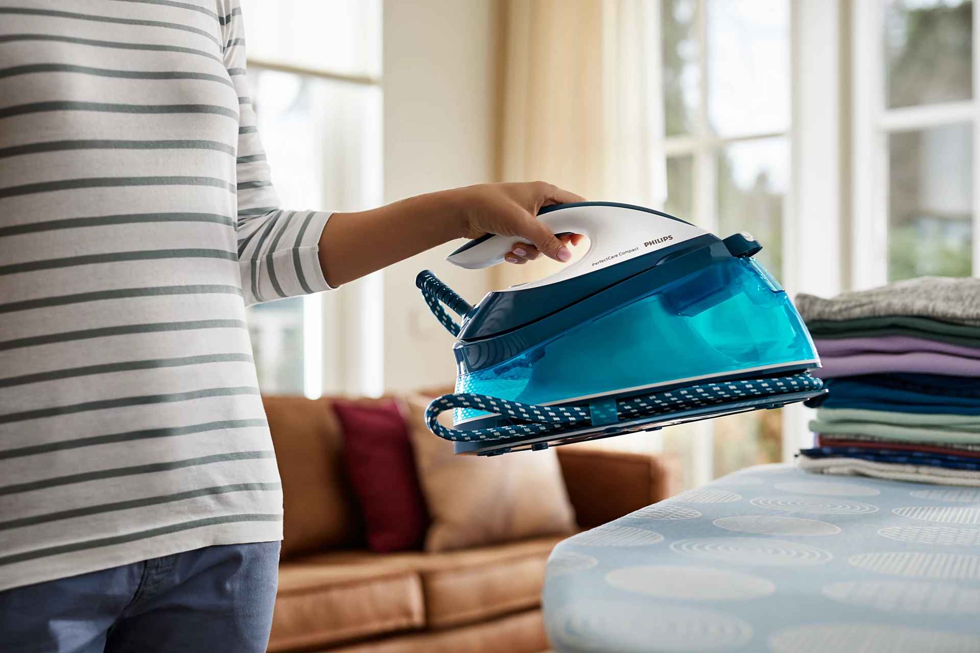 Roka paceļ Philips tvaika gludināšanas sistēmu PerfectCare Compact, zili un balti. Gludinātās veļas kaudze ir novietota sānos, uzsverot profesionālus rezultātus.