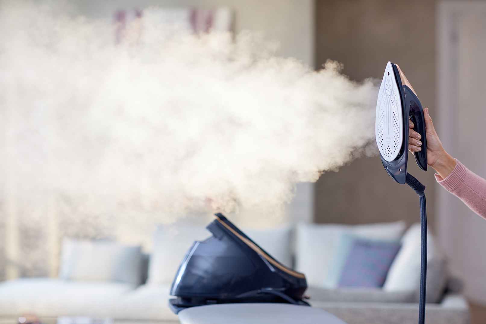 Philips gudināšanas sistēmasi: augstākās klases apģērbu kopšana, kas izdala jaudīgu tvaiku. Lietotājs tur moderno gludekli ar gludināšanas virsmu uz priekšu. Redzama pamatdaļa; attēlota luksusa dzīves atmosfēra.