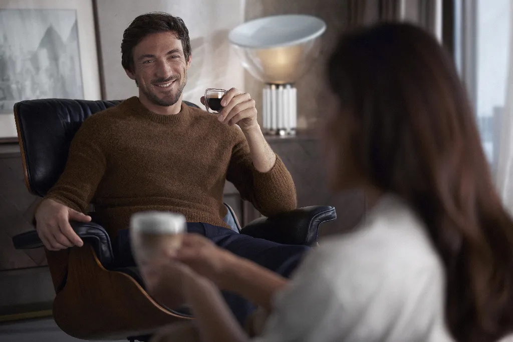 12 copy Vīrietis ērti sēž atzveltnes krēslā, smaida, turot rokās espreso, bet cits cilvēks priekšplānā piedāvā kafiju ar piena putām.