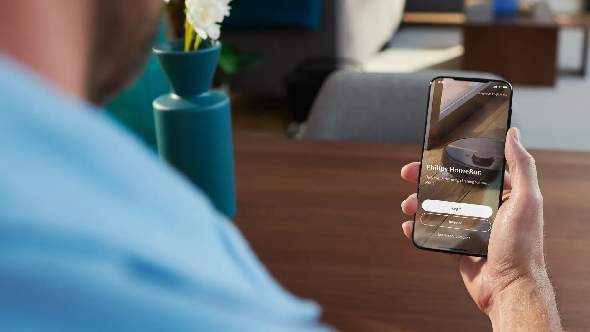 Vīrietis tur rokās tālruni, kurā redzama Philips HomeRun lietotnes saskarne. Attēlots Philips robotizētais putekļsūcējs, kas sola ikdienas mitro un sauso dziļo tīrīšanu, kā arī iespējas pieteikties, reģistrēties vai lietot ierīci bez konta.