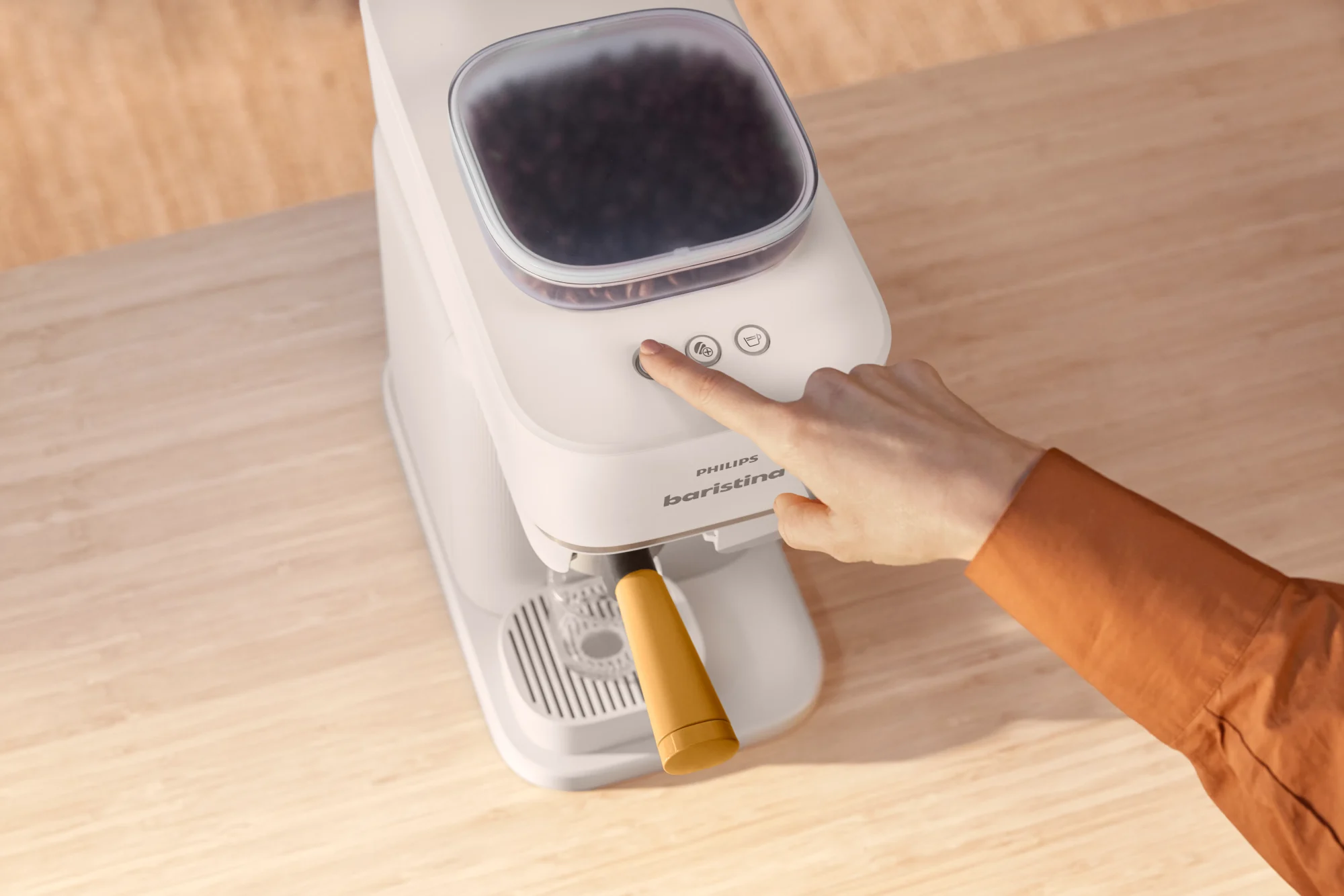 Roka, kas nospiež Philips Baristina kafijas automāta palaišanas pogu, ierīcē redzamas kafijas pupiņas. Izbaudi no veselām pupiņām pagatavotu augstākās kvalitātes kafiju. Baristina ierīce novietota uz gaiša koka darba virsmas.