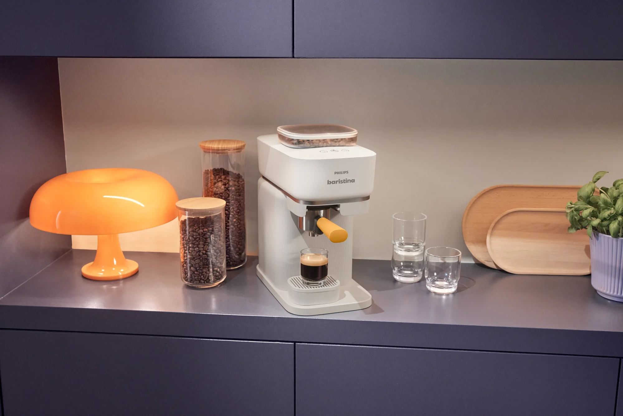 Stilīgs Philips Baristina espreso automāts atrodas uz galda blakus traukiem ar kafijas pupiņām un svaigi pagatavotai espresso tasei. Moderns virtuves iekārtojums.